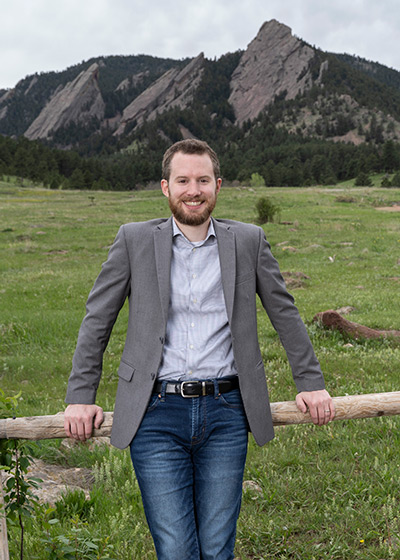 Ron Stauffer smiles while posing in front of the Flatirons in Boulder. He's wearing blue jeans and a gray sport coat and smiling.